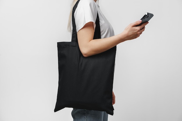 Foto imagen recortada de una mujer con una bolsa de compras de algodón negro usando un teléfono inteligente con fondo blanco