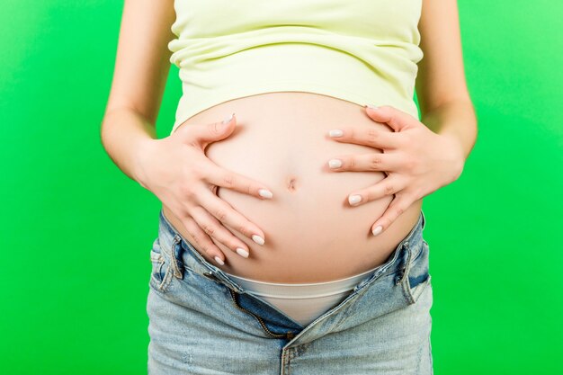 Imagen recortada de una mujer embarazada en jeans desabrochados que muestra la panza de su bebé en una superficie colorida con espacio de copia