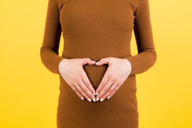 Imagen recortada de mujer embarazada en vestido marrón haciendo forma de corazón con las manos sobre su abdomen en fondo amarillo. Concepto de golpe de bebé. Copie el espacio.