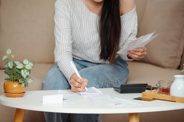 Foto imagen recortada de una mujer joven haciendo contabilidad en casa, comprobando facturas y escribiendo en un cuaderno