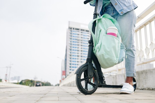 Imagen recortada de mujer joven con mochila de pie en scooter eléctrico con una taza de bebida en el bolsillo