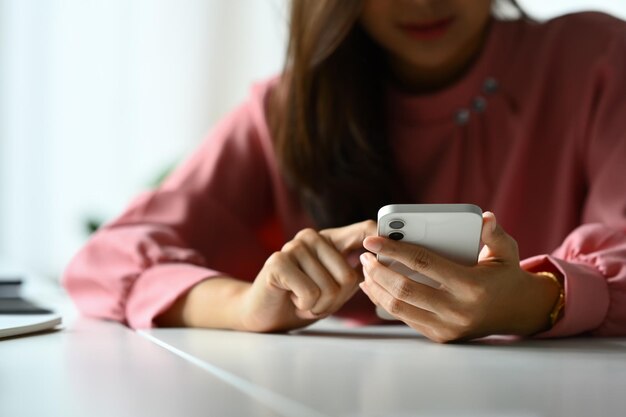 Foto imagen recortada de una mujer joven usando un teléfono inteligente en el escritorio de la oficina revisando las redes sociales comunicándose en línea