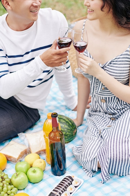Imagen recortada de la sonriente joven pareja asiática brindando con copas de vino tinto al tener un picnic romántico en el día de San Valentín