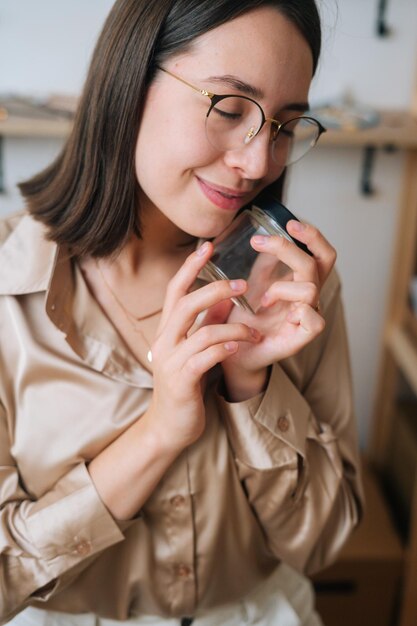 Imagen recortada vertical de una joven feliz con gafas sosteniendo un pequeño frasco de vidrio transparente vacío en las manos sentada en la mesa, con los ojos cerrados. Proceso de elaboración de velas naturales artesanales en taller.