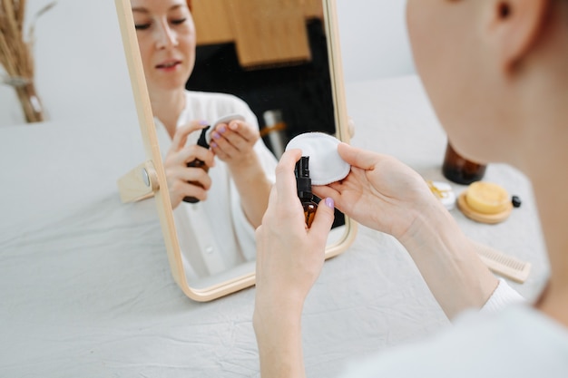 Imagen de reflejo de una mujer sentada frente al espejo, aplicando agua micelar. Usar productos y cosas ecológicos.