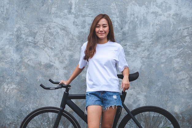 Imagen de retrato de una hermosa joven asiática con camiseta blanca y pantalón corto de jean con bicicleta y fondo de pared de hormigón