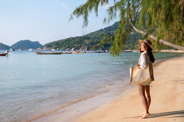 Imagen de retrato de una hermosa joven asiática con sombrero y bolso paseando por la playa