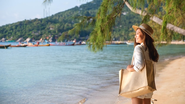 Imagen de retrato de una hermosa joven asiática con sombrero y bolso paseando por la playa