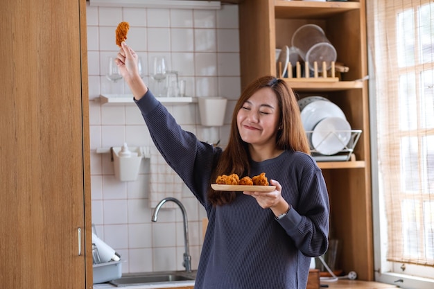Imagen de retrato de una hermosa joven asiática sosteniendo pollo frito y bailando en la cocina en casa