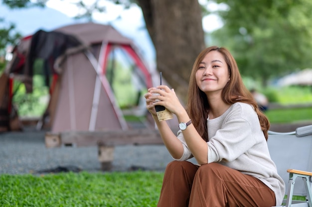 Imagen de retrato de una hermosa joven asiática tomando café mientras se sienta en una silla de camping con una carpa en el parque