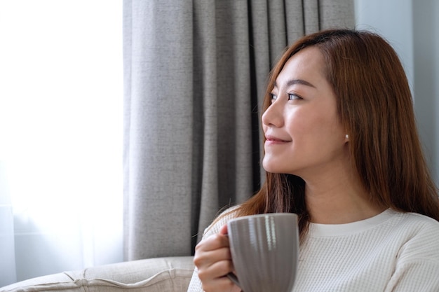 Imagen de retrato de una hermosa mujer asiática mirando por la ventana mientras bebe café caliente y se relaja en un sofá en el dormitorio en casa
