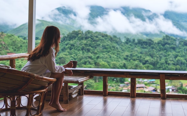 Imagen de retrato de una hermosa mujer asiática sosteniendo y bebiendo café caliente, sentada en el balcón y mirando las montañas y la naturaleza verde