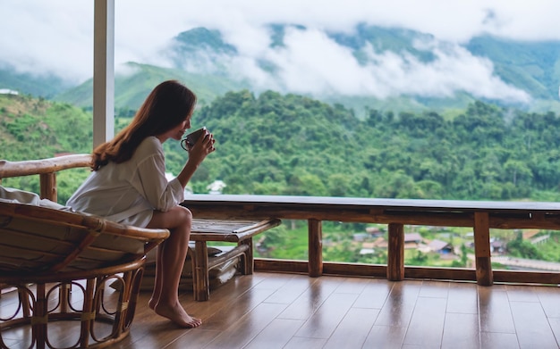 Imagen de retrato de una hermosa mujer asiática sosteniendo y bebiendo café caliente, sentada en el balcón y mirando las montañas y la naturaleza verde