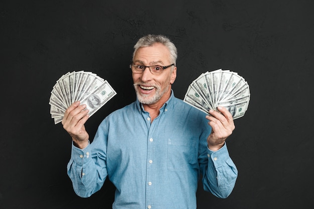 Imagen del rico hombre adulto feliz de los años 60 con canas sosteniendo dinero dos fanáticos de billetes de 100 dólares y regocijándose de su riqueza, aislado sobre la pared negra