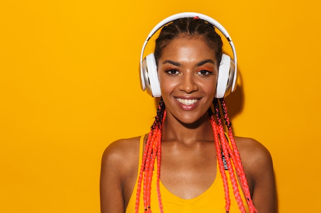 Imagen de seductora mujer afroamericana con trenzas afro sonriendo y escuchando música con auriculares aislado sobre pared amarilla