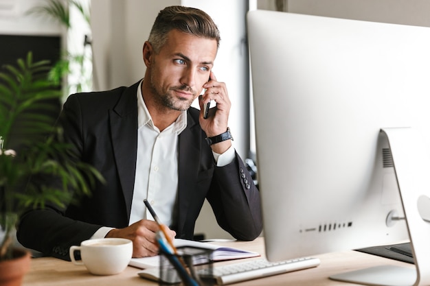 Imagen del serio empresario de 30 años vistiendo traje hablando por teléfono celular mientras trabaja en la computadora en la oficina