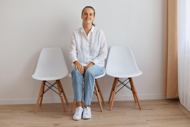 Imagen de sonriente mujer adulta joven atractiva satisfecha sentada en una silla contra la pared blanca, vestida con camisa y jeans, mirando a la cámara con expresión facial tranquila y optimista.