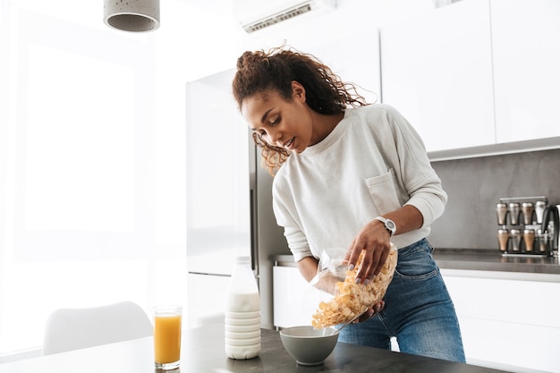 Imagen de sonriente mujer afroamericana haciendo el desayuno con copos de maíz y leche, en cocina elegante