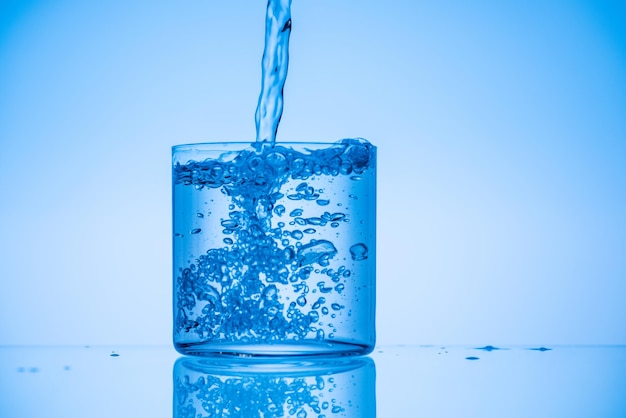Imagen tonificada de agua que se vierte en un vaso lleno sobre fondo azul.