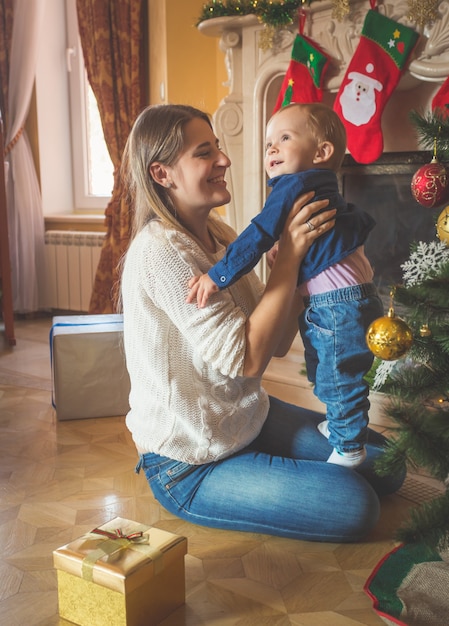 Imagen de tonos de feliz madre sonriente y su hijo de 1 año posando en el árbol de Navidad