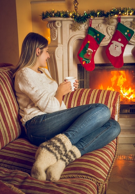 Imagen de tonos de joven bella mujer en calcetines de lana relajándose en el sofá en la chimenea y bebiendo té