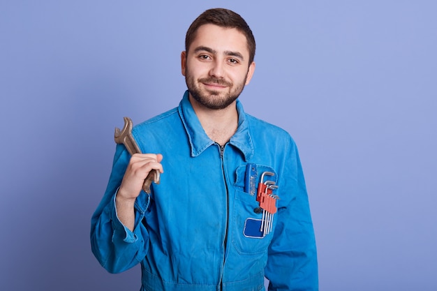 Foto imagen del trabajador de la construcción trabajador encantado mirando directamente a la cámara, de pie aislado sobre fondo lila en estudio, sosteniendo la llave en una mano. concepto de personas y lugar de trabajo.