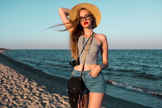 Foto imagen de verano al aire libre de hermosa mujer rubia con sombrero de paja caminando cerca del mar.