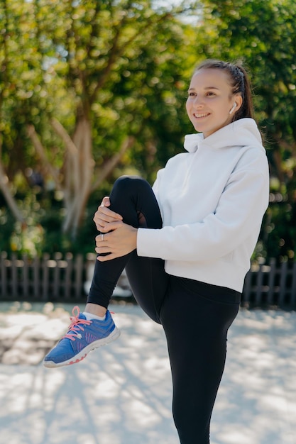 Imagen vertical de una mujer enérgica que lleva un estilo de vida activo mantiene ejercicios de rodilla levantados al aire libre vestidos con ropa activa levanta las sonrisas de las piernas escucha agradablemente la pista de audio en los auriculares inalámbricos Entrenamiento