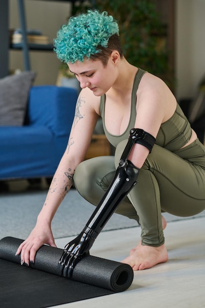 Imagen vertical de mujer joven con brazo protésico preparando colchoneta de ejercicio para entrenamiento deportivo en casa