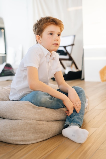 Imagen vertical de un niño confiado mirando a la cámara sentado en una almohada en el suelo en la sala de estar con un interior moderno y luminoso