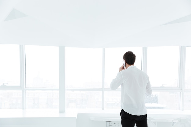 Imagen de vista posterior del hombre vestido con camisa blanca hablando por teléfono cerca de una gran ventana blanca.