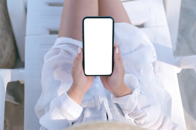 Imagen de la vista superior de la mano de una mujer sosteniendo un teléfono móvil blanco con pantalla de escritorio en blanco mientras se acuesta en una silla de playa