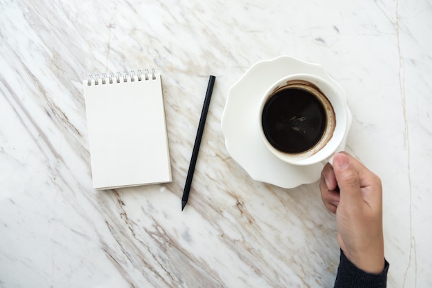 Imagen de la vista superior de una mano sosteniendo la taza de café con un cuaderno blanco en blanco en la mesa