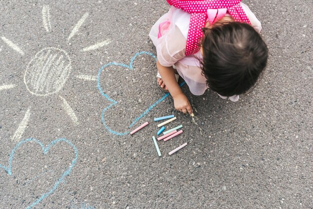 Imagen de la vista superior de una niña feliz que lleva un vestido rosa y una mochila dibujando con tizas de colores en la acera Lindo niño en edad preescolar juega al aire libre en el pavimento Actividad educativa para niños pequeños afuera