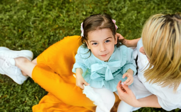 Imagen de la vista superior de un niño feliz jugando con su madre disfrutando del tiempo juntos al aire libre Niña alegre comiendo algodón de azúcar con su madre sentada en la hierba verde en el parque Día de la madre