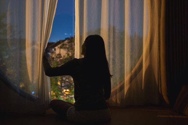 Imagen de la vista trasera de una mujer abriendo la cortina del dormitorio y mirando por la ventana por la noche