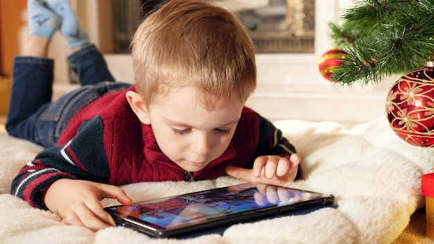 Imágenes de primer plano 4k de niño sonriente jugando juegos en tableta digital mientras está acostado bajo el árbol de Navidad en casa. Niño divirtiéndose en celebraciones y vacaciones de invierno.