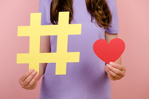 Imágenes recortadas de una mujer joven que viste una camiseta muestra un gran letrero de hashtag amarillo grande y un pequeño corazón rojo, posando aislado sobre fondo rosa en el estudio, símbolo de la caridad en Internet