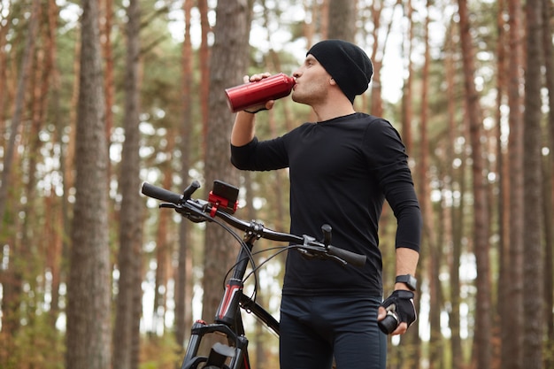 Imagens ao ar livre horizontal de bom ajuste esportista beber água de garrafa vermelha, tendo uma curta parada, andando de bicicleta na floresta sozinha, aderindo ao estilo de vida saudável.