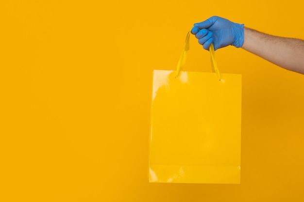 Imagens de close up do conceito de entrega. Mão com luvas azuis segurando grande bolsa colorida isolada sobre o fundo amarelo.