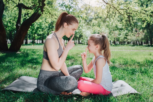 Imagens engraçadas as meninas sentadas no carimate fora no parque e mostrando o símbolo do silêncio para o outro. Eles estão sorrindo e rindo um pouco. Conceito de ioga e pilates.