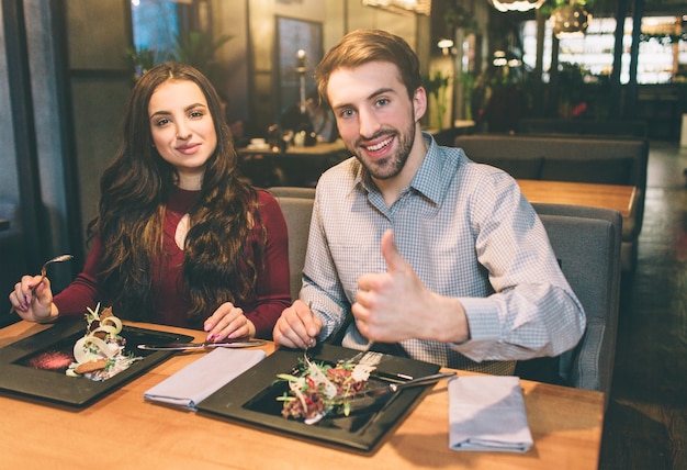 Foto imagens publicitárias de homem e mulher sentada à mesa com um pouco de comida. eles estão olhando para a câmera e sorrindo. homem está mostrando o polegar grande