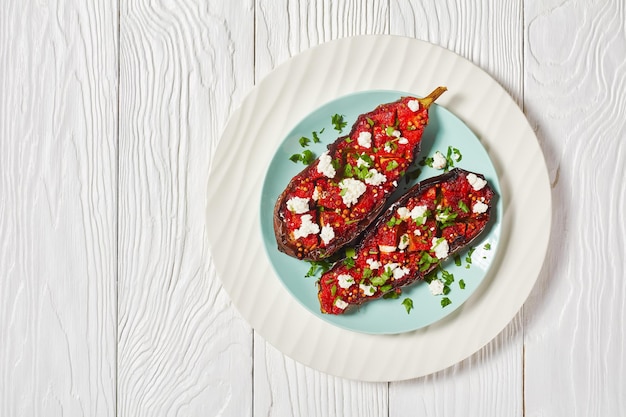 Imam Bayildi con queso feta desmenuzado, berenjenas rellenas de sabrosos tomates, cebollas y especias en un plato sobre una mesa de madera blanca, espacio libre