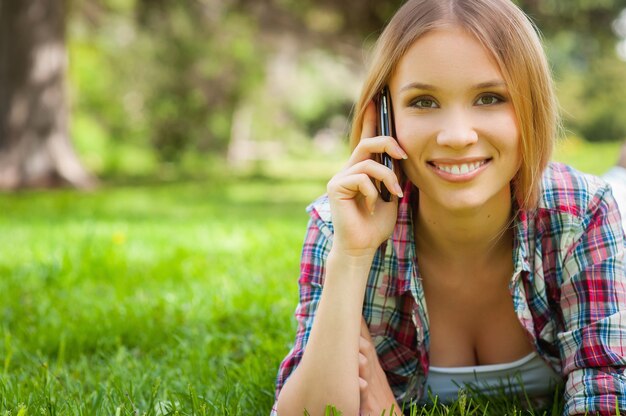 Immer in Kontakt. Schöne junge Frau, die mit dem Handy spricht und lächelt, während sie auf dem Gras im Park liegt