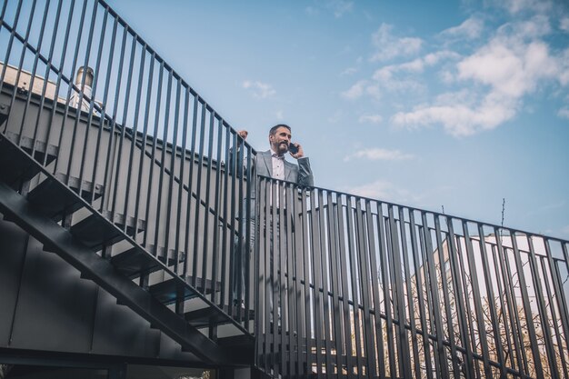 Immer in Kontakt. Selbstbewusster bärtiger Geschäftsmann im Anzug, der auf Smartphone kommuniziert, das auf Stufen der Treppe draußen steht