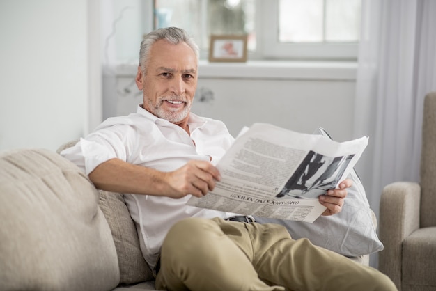 Immer lächeln. Fröhlicher Mann, der Lächeln auf seinem Gesicht hält und Zeitung hält, während er gerade in die Kamera schaut