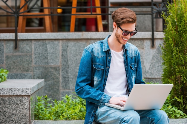 Immer verbunden. Lächelnder junger Mann, der am Laptop arbeitet, während er im Freien sitzt