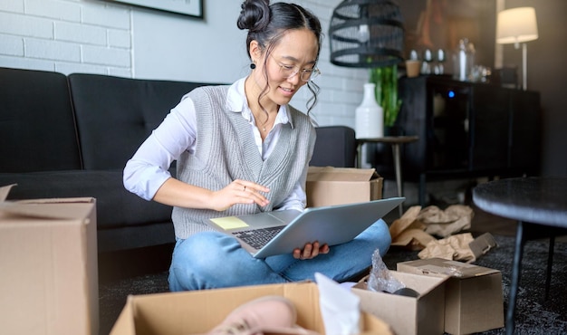 Immobilien-Laptop und asiatische Frau, die in ein neues Zuhause ziehen, während sie Hypotheken online planen oder berechnen Umzugsboxen Computer und weibliche Eigentümer oder Fernarbeiter im Wohnzimmer des Hauses