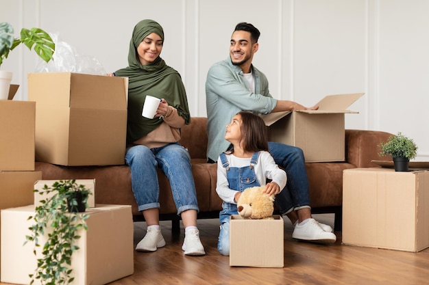 Immobilienkauf, Hausumzug, Umzugskonzept. Porträt einer fröhlichen muslimischen Familie, die Kartons auspackt, mit offenen Paketen auf der Couch und dem Boden im Wohnzimmer sitzt. Gehäuse