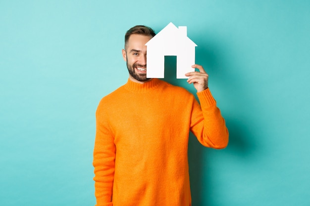 Immobilienkonzept. Glücklicher junger Mann, der nach Hausmiete sucht, Hauspapiermaket hält und lächelt, über blauem Hintergrund stehend.